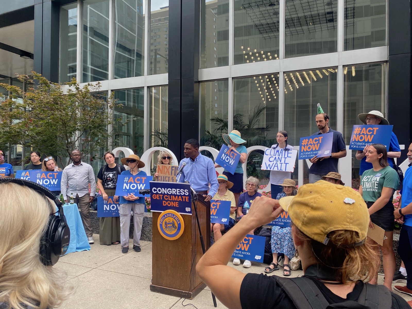 Christopher Johnson, a regional representative for U.S. Senator Bob Casey, stands speaking at a podium in front of a crowd of people holding signs that say "Act Now on Climate"