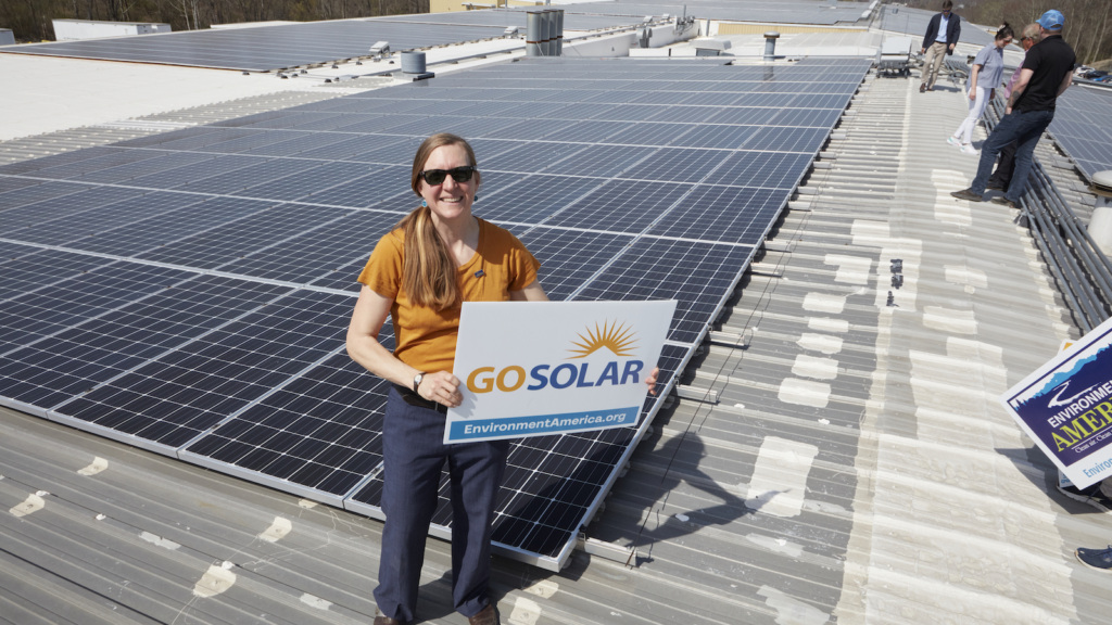 Environment America 100% Renewable Energy Senior Director Johanna Neumann stands on a warehouse rooftop covered in solar panels holding a sign that reads "go solar."